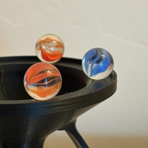 May include: Three glass marbles with swirling orange, black, and blue patterns rest in a black, 3D-printed holder. The marbles are clear, allowing the colorful designs to be visible. The holder has a unique, modern design.