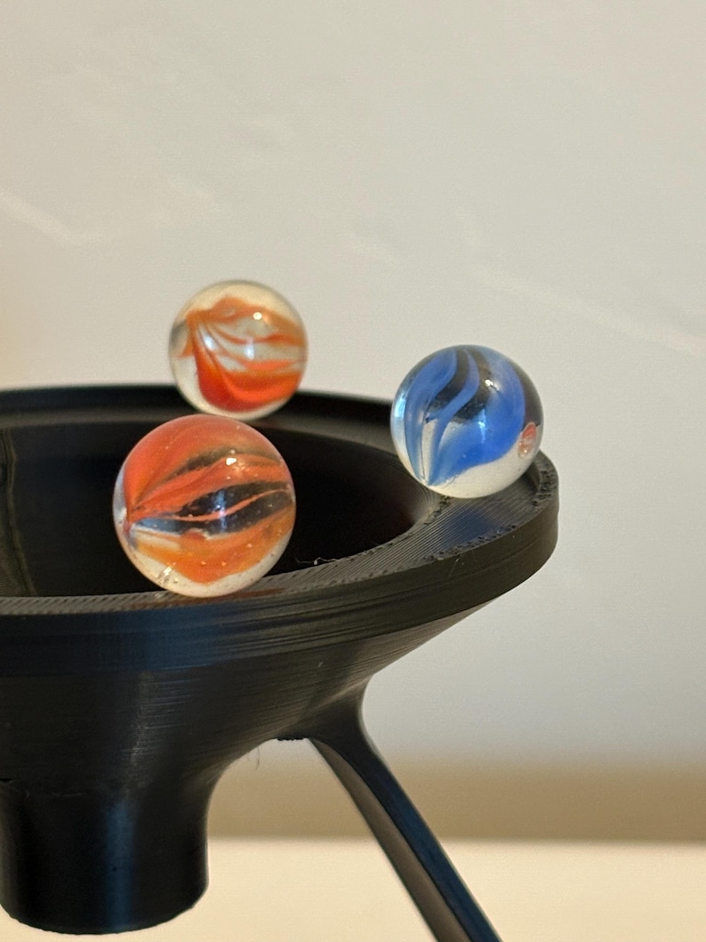 May include: Three glass marbles with swirling orange, black, and blue patterns rest in a black, 3D-printed holder. The marbles are clear, allowing the colorful designs to be visible. The holder has a unique, modern design.
