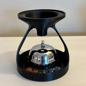 May include: A black, 3D-printed bell stand with a chrome bell and colorful marbles. The stand has a unique, modern design with a circular base and a flared top. The bell is centered above the marbles.