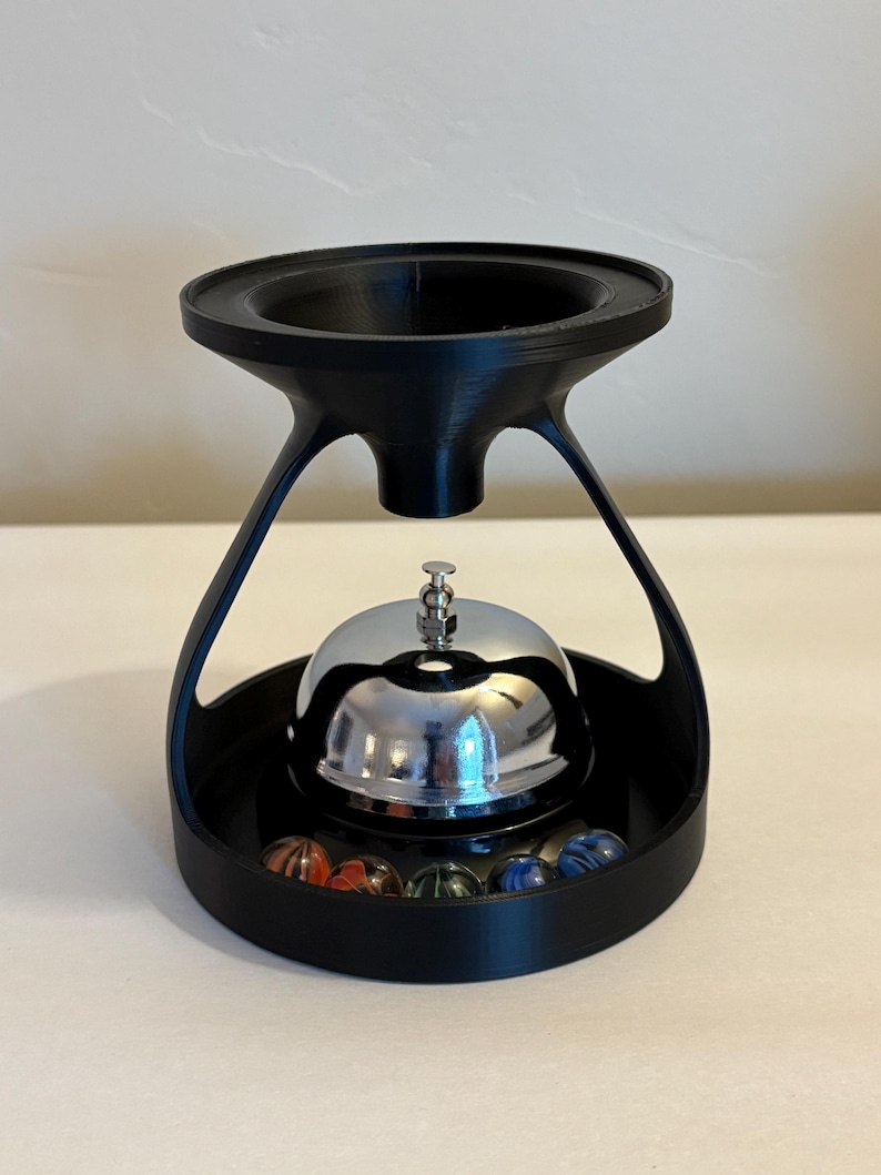 May include: A black, 3D-printed bell stand with a chrome bell and colorful marbles. The stand has a unique, modern design with a circular base and a flared top. The bell is centered above the marbles.