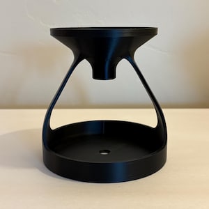 May include: A black, hourglass-shaped object with a wide, circular base and a flared top. The top and base are connected by curved supports, creating an open, airy design. The object is made of a solid, matte material.
