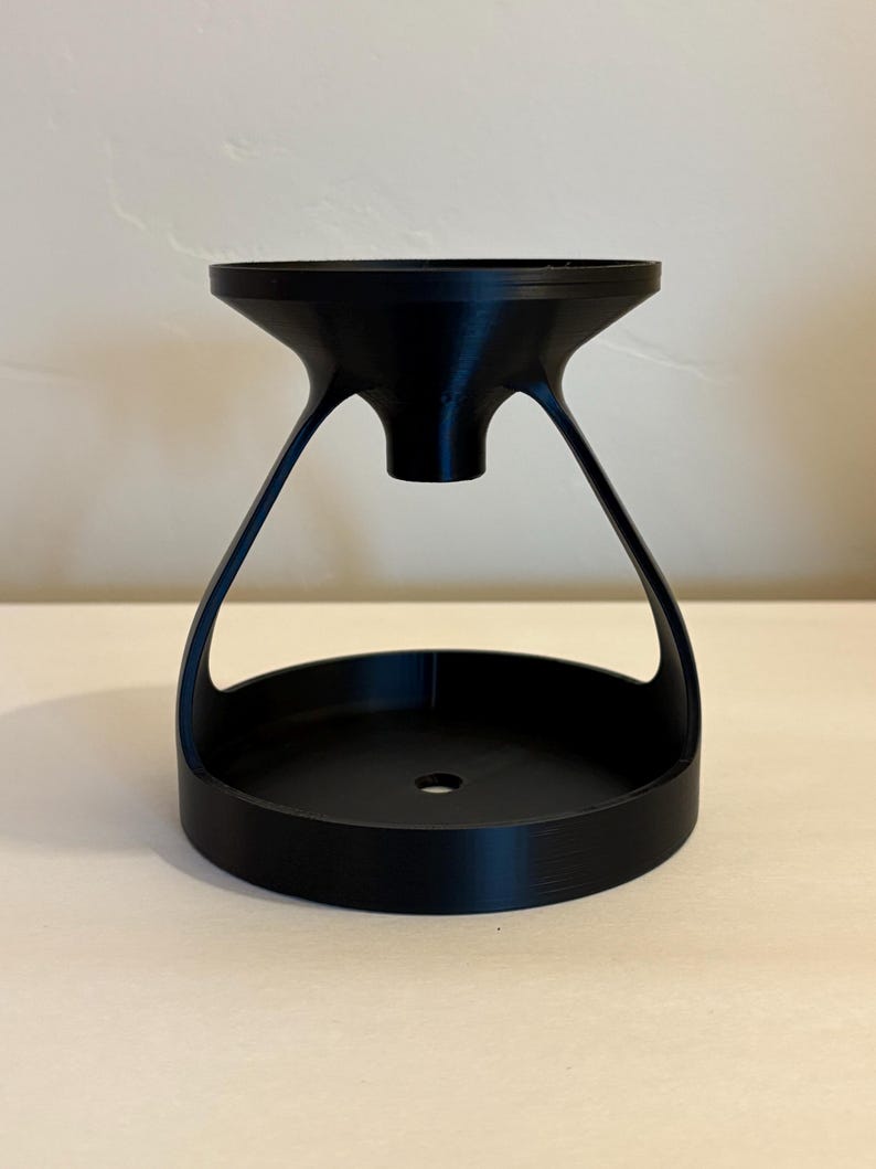 May include: A black, hourglass-shaped object with a wide, circular base and a flared top. The top and base are connected by curved supports, creating an open, airy design. The object is made of a solid, matte material.