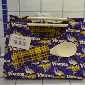 May include: Purple and gold quilted fabric utensil holder with the Minnesota Vikings logo. The holder has two wooden dowels for hanging utensils and a pocket for a wooden spoon.