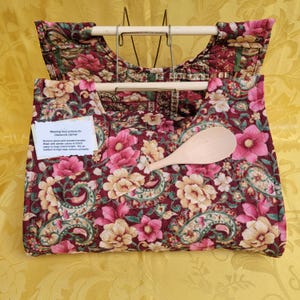May include: A quilted casserole carrier featuring a dark red floral and paisley pattern with two light wood handles. A light wood serving spoon is tucked into the front. A white tag with black text provides washing instructions for the carrier.