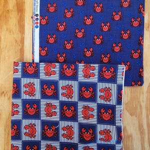 May include: Two pieces of blue fabric with nautical patterns. One features red crabs and blue anchors on a solid background. The other has red crabs in white-lined blue squares. The fabric is from "Fabric & Craft Stores".