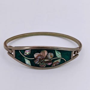May include: A silver-toned metal hinged bangle bracelet featuring a central curved panel. The panel displays a dark green background inlaid with iridescent abalone shell pieces arranged in the shape of a flower with leaves and thin silver stems.