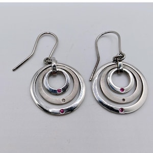 May include: A pair of silver-toned earrings with a circular design. Each earring features three concentric circles, with small red gemstones set at intervals. The earrings have a simple hook closure.