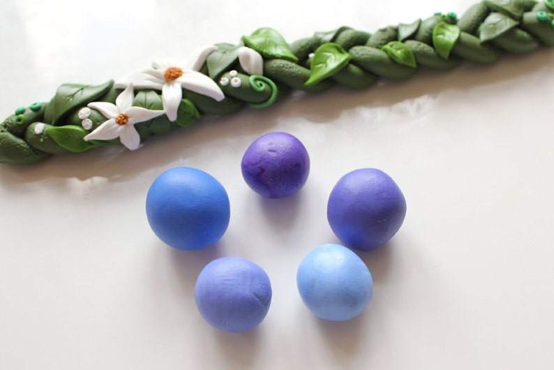 May include: Five balls of clay in different shades of blue and purple. The balls are arranged in a circle on a white surface.