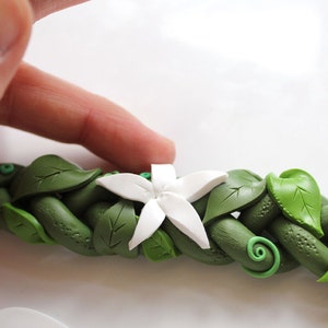May include: A white polymer clay flower with five petals is placed on top of a green polymer clay vine with leaves. The vine is braided and has a textured surface.