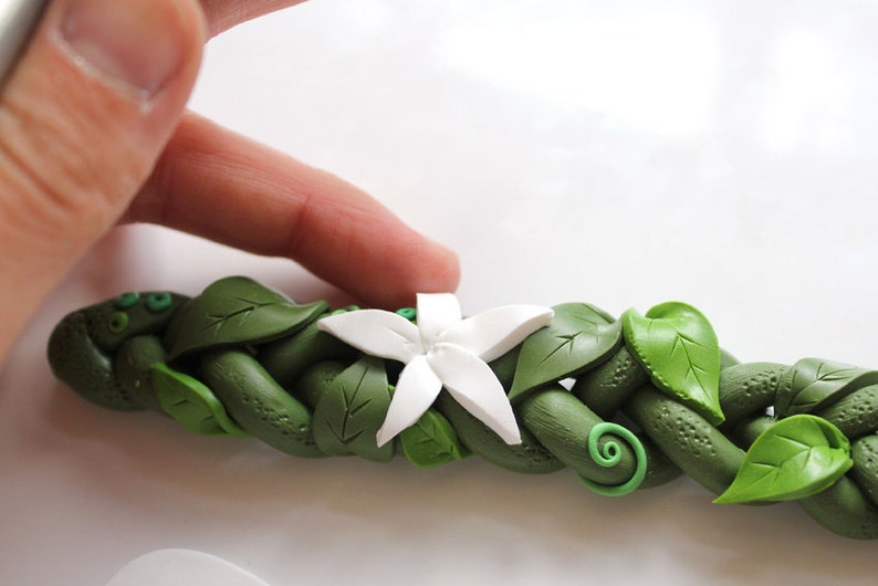 May include: A white polymer clay flower with five petals is placed on top of a green polymer clay vine with leaves. The vine is braided and has a textured surface.