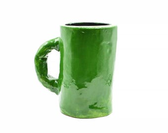 May include: A handmade, glossy green ceramic mug with a rounded handle. The mug has a cylindrical shape and a dark interior. The mug is a vibrant shade of green and appears to be hand-glazed.