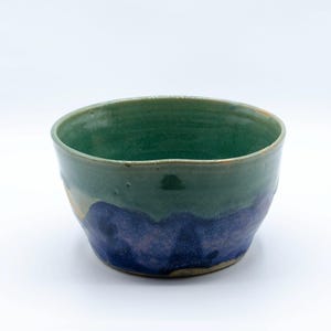 May include: A handmade ceramic bowl with a green upper section and a blue lower section. The bowl has a slightly irregular shape and a rustic, artisanal appearance. The interior is a lighter shade of green.