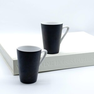 以下が含まれることがあります： 白と黒のセラミックマグカップ2つ。内側とハンドルは白で、円筒形でわずかに先細りになっています。白い表面、おそらく本の上に置かれ、背景は白です。マグカップはミニマリストデザインです。