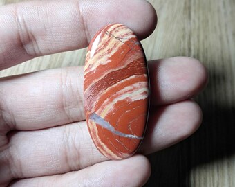 May include: An oval-shaped polished gemstone cabochon featuring a vibrant pattern of swirling terracotta red, cream, and dark brown bands. The stone has a smooth, glossy surface with a distinct thin white mineral vein running across the lower section.