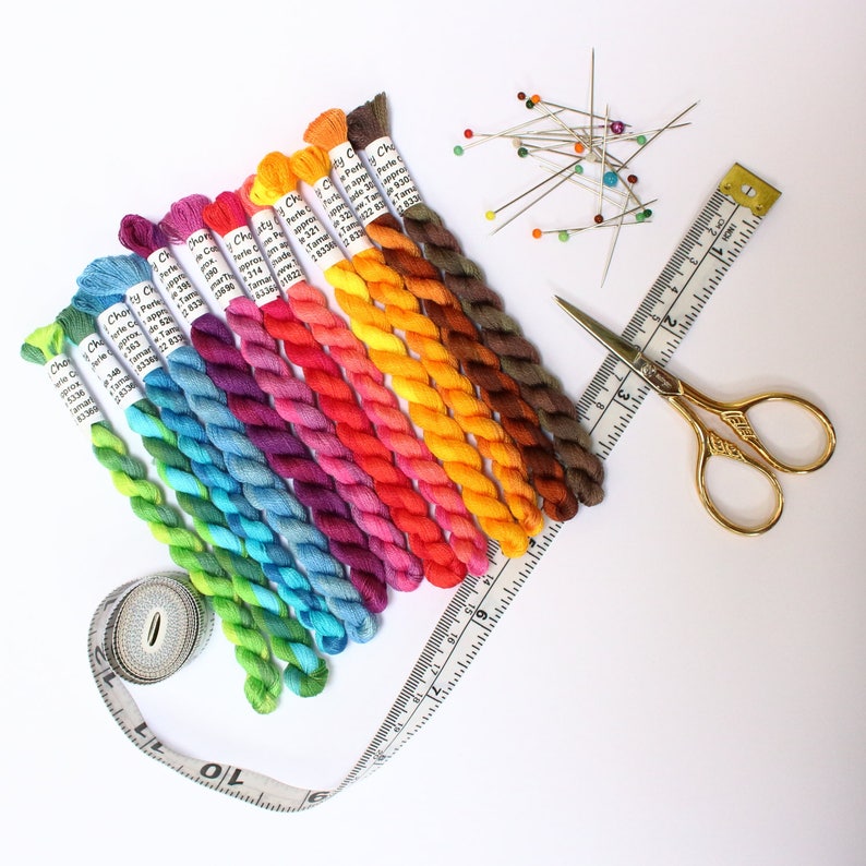 May include: A selection of vibrant embroidery threads in a rainbow spectrum, alongside sewing accessories. Features a measuring tape, gold scissors, and pins with coloured heads. The threads are labelled with brand details and colour codes.