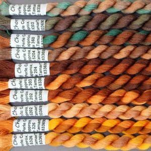 May include: A selection of embroidery threads in shades of brown, orange, and green. Each skein is labelled with the brand name and colour code. The threads are tightly wound, ready for use in needlework projects.
