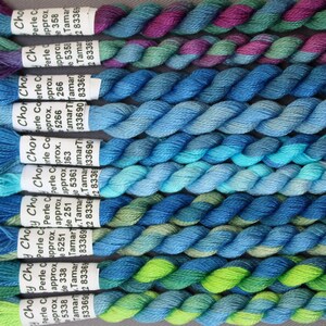 May include: A close-up shot of several skeins of Choy Choy Perle Cotton thread in shades of blue, green, and purple. Each skein is labelled with the brand name and product details. The threads are tightly twisted, displaying a range of colours.