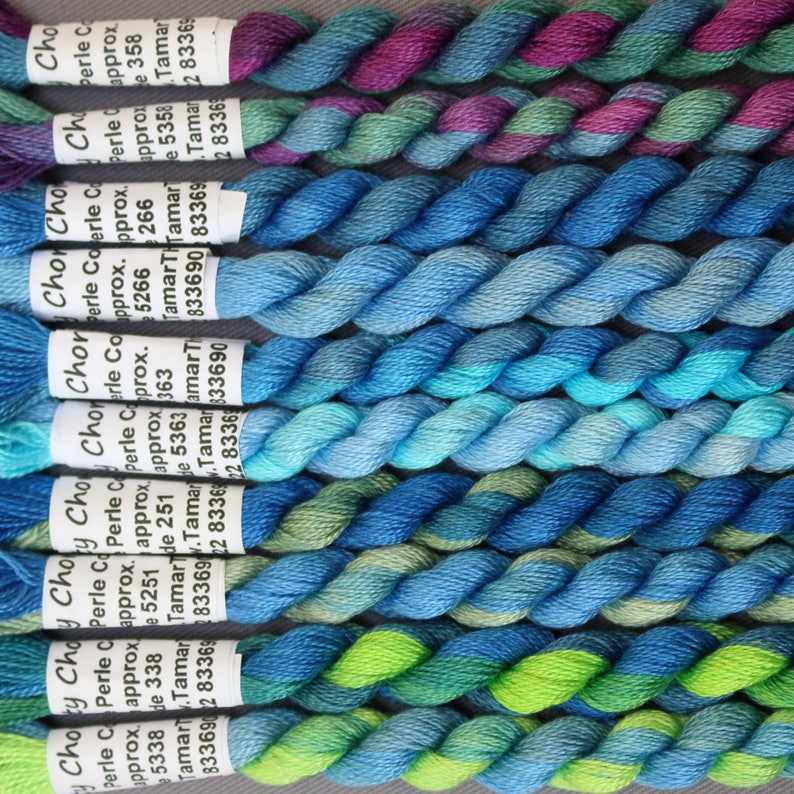 May include: A close-up shot of several skeins of Choy Choy Perle Cotton thread in shades of blue, green, and purple. Each skein is labelled with the brand name and product details. The threads are tightly twisted, displaying a range of colours.