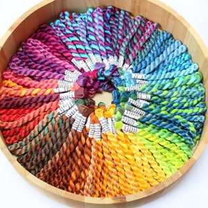 May include: A wooden bowl containing a colourful selection of embroidery floss, arranged in a circular pattern. The floss displays a spectrum of colours, including purple, pink, blue, green, yellow, orange, and brown. Each skein has a white label attached.