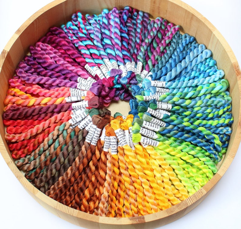 May include: A wooden bowl containing a colourful selection of embroidery floss, arranged in a circular pattern. The floss displays a spectrum of colours, including purple, pink, blue, green, yellow, orange, and brown. Each skein has a white label attached.