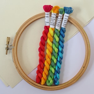 May include: A wooden embroidery hoop with a canvas and a selection of colourful embroidery floss. The floss is arranged in a rainbow gradient, with shades of red, orange, yellow, green, and blue. The floss labels are visible.