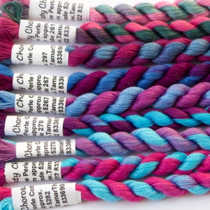 May include: A selection of colourful embroidery floss skeins, tightly twisted and displaying a range of hues. The floss features shades of pink, purple, blue, and teal. Each skein has a white label with text. Suitable for cross-stitch and embroidery.