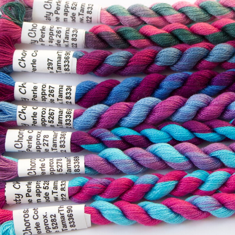 May include: A selection of colourful embroidery floss skeins, tightly twisted and displaying a range of hues. The floss features shades of pink, purple, blue, and teal. Each skein has a white label with text. Suitable for cross-stitch and embroidery.