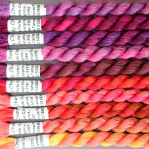 May include: A selection of embroidery threads in various shades of pink, red, and orange. The spools are arranged in rows, displaying a gradient of colours. Each spool has a label with text.