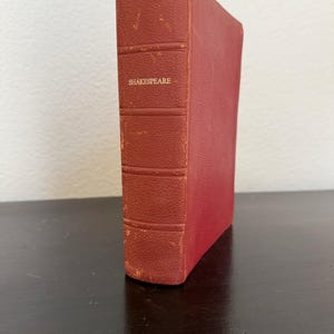 May include: A vintage hardcover book with a textured red cover. The spine features horizontal raised bands and the word SHAKESPEARE printed in gold lettering. The edges of the spine show signs of wear and fading, revealing a lighter red tone.