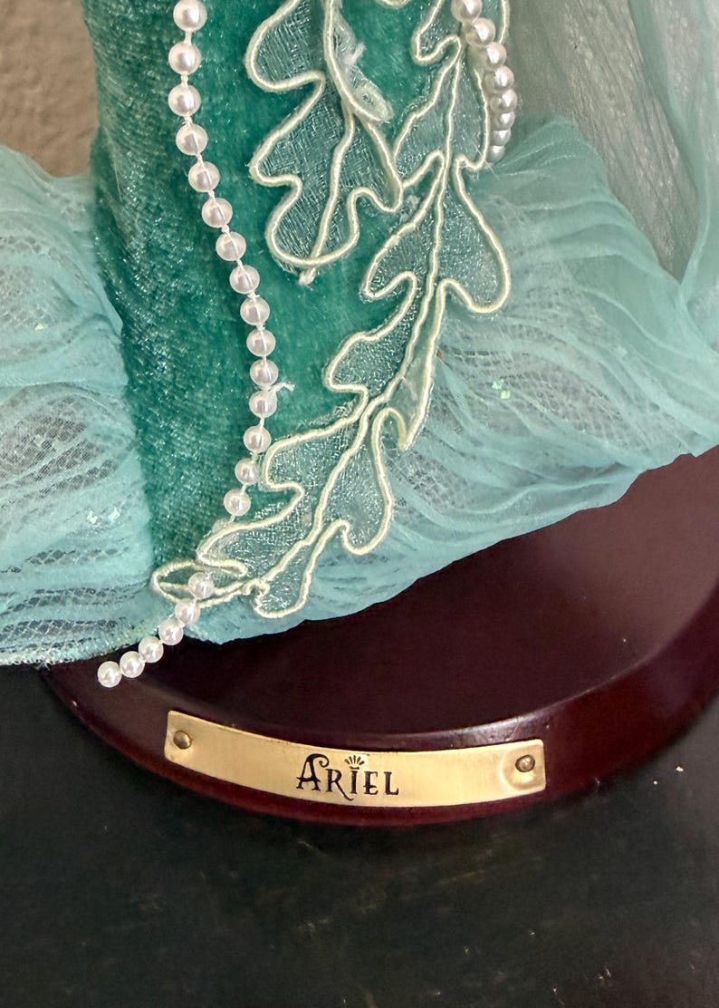 Może przedstawiać: Turkusowa figurka z perłowym naszyjnikiem i dekoracyjnym wzorem liści. Figurka ma złotą tabliczkę z napisem "ARIEL". Podstawa jest w kolorze ciemnobrązowym.