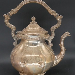 May include: Silver teapot with a curved handle and decorative detailing. The teapot features a lid with a floral finial and a spout with an ornate design. The body of the teapot is rounded with vertical ridges and three small, round knobs.