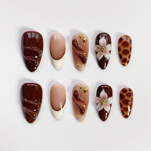 May include: A collection of ten artificial nails in various designs. The nails feature shades of brown, beige, and white. Some nails have a swirl design, gold accents, a floral design, and a giraffe print.