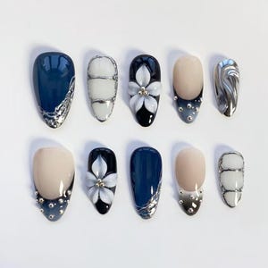 May include: A set of ten artificial nails in various designs. The nails feature a range of colors, including navy blue, black, and nude, with silver and white accents. Some nails have floral embellishments or a metallic swirl design.