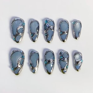 May include: A collection of ten false nails, each featuring a silver metal border and a light blue central design. Small, blue gemstones are set into each nail. The nails are displayed on a white background.