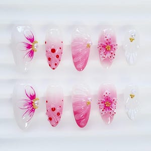 May include: A set of ten artificial nails with a floral design. The nails feature a white base with pink and red accents, including flowers and dots. Some nails have gold embellishments.