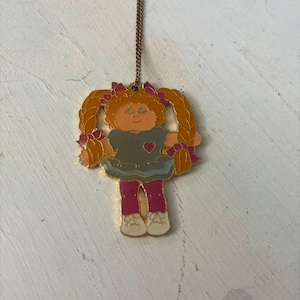 Può includere: Un ciondolo in metallo dorato su una catenina sottile con un'illustrazione smaltata di una bambola con trecce arancioni, che indossa un vestito grigio con un piccolo cuore rosso, leggings magenta e scarpe bianche, con fiocchi rosa.