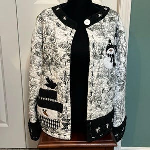 May include: A quilted jacket featuring a black and white winter forest scene with snowman appliqués. The jacket has a round neckline, a single white button closure, and black cuffs with white embroidered snowflake details throughout.