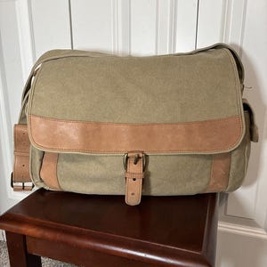 Op de afbeelding: Een olijfgroene canvas messenger bag met een bruin lederen accent over de voorkant en op de riemen. De tas heeft een klepsluiting met een gesp en staat op een donkerbruine houten kruk.