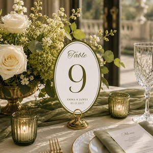 May include: An oval table number sign featuring the text Table 9, Emma & James, and 03.11.2027 in dark green on a cream background with a dark green border. The sign is held by a gold metal stand and is placed among white roses and greenery.