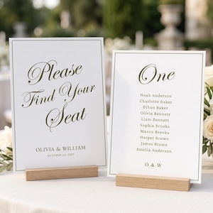 May include: Two white rectangular wedding seating chart signs with dark olive green text and thin borders, displayed on light wood stands. The left sign reads Please Find Your Seat, Olivia & William, October 20, 2027. The right sign lists names under One.