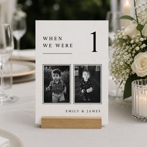 Könnte beinhalten: Eine weiße, rechteckige Tischnummernkarte mit dem Text "WHEN WE WERE" und der Zahl "1" in schwarzer Serifenschrift. Die Karte zeigt zwei Schwarz-Weiß-Fotos eines Kindes und die Namen "EMILY & JAMES" am unteren Rand, gehalten von einem Holzblock.