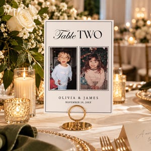 May include: A rectangular white table number card featuring the text Table TWO, two square photographs of children, and the names OLIVIA & JAMES with the date NOVEMBER 20, 20XX. The card is held upright by a circular gold metal stand on a table.
