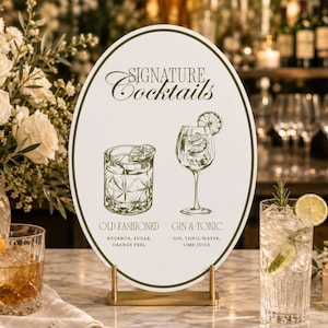 May include: An oval white sign with a dark green border and gold stand, featuring the text "SIGNATURE COCKTAILS" and illustrations of an Old Fashioned and a Gin & Tonic with their respective ingredients listed in dark green lettering.