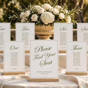 May include: A collection of white wedding seating chart cards with dark green text, each held by a light wood stand. The central sign reads Please Find Your Seat, Olivia & William, October 20, 2027. Other cards display table numbers and guest names.