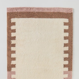 May include: Rectangular bath mat with a cream-colored center and a border featuring a brown and pink color scheme. The border has a stepped design. The mat is made of a soft, plush material, suitable for a bathroom setting.