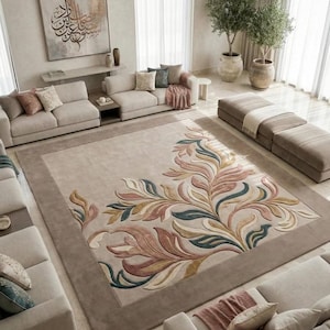 May include: A large beige area rug with a floral design in shades of pink, blue, and yellow. The rug has a brown border and is in a living room with beige sofas, cushions, and potted plants. The room has a neutral colour palette.