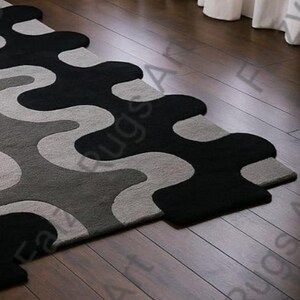 May include: A modern area rug with a unique, flowing design. The rug features a pattern of black, gray, and light gray organic shapes. The rug is placed on a dark wood floor, adding a touch of contemporary style to any room.