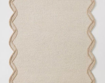 May include: A rectangular beige rug with a textured surface and a scalloped border in a lighter beige color. The rug has a simple, neutral design, suitable for various interior styles.