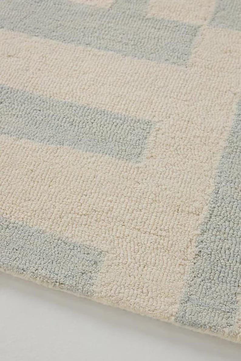 May include: Close-up of a textured rug with a geometric pattern. The rug features alternating bands of light blue and off-white, creating a soft, inviting aesthetic. The fibers appear plush and comfortable.