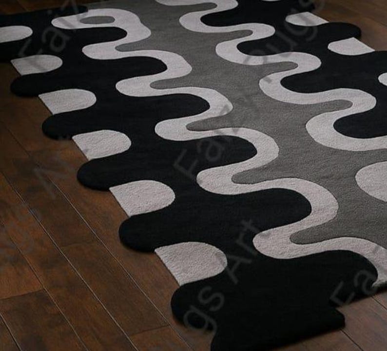 May include: A modern area rug with a wavy pattern in black, gray, and off-white. The rug's design features large, flowing shapes that create a bold visual statement. The rug is placed on a wooden floor.